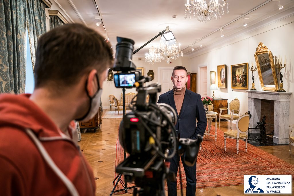 operator kamery w okienku obserwuje scenę z drugiego planu - aktora na tle historycznego wnętrza