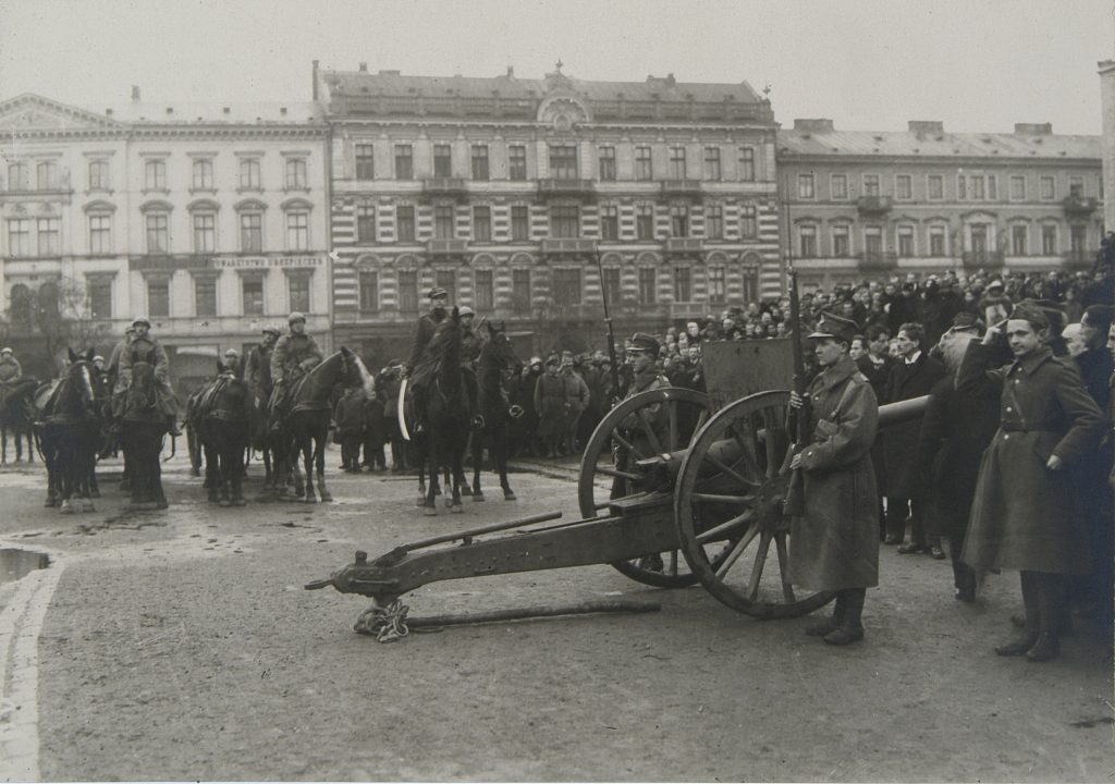 czarno-białe zdjęcie olacu. na pierwszym planie działo, obok zgromadzony tłum i żolnierze na koniach, w tle kamienice