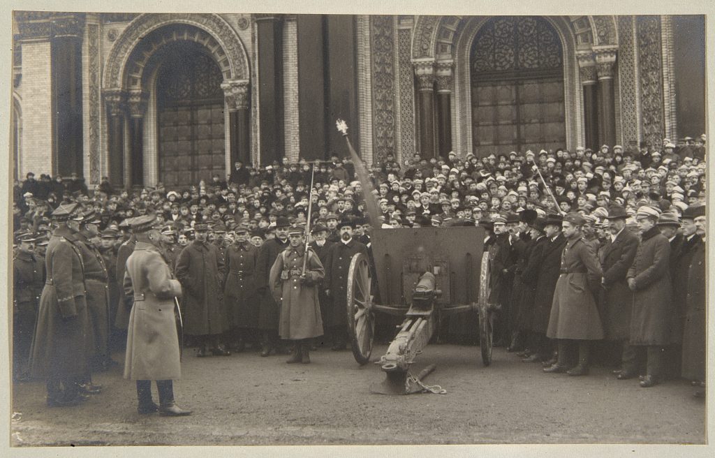 tłum ludzi za stojącym w centrum czarno-białej fotografii działem, w tle ozdobny budynek