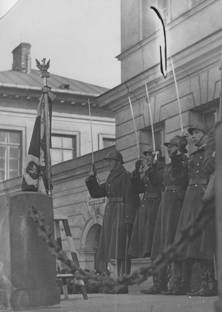 grupa żołnierzy salutujących szablami, w tle budynek