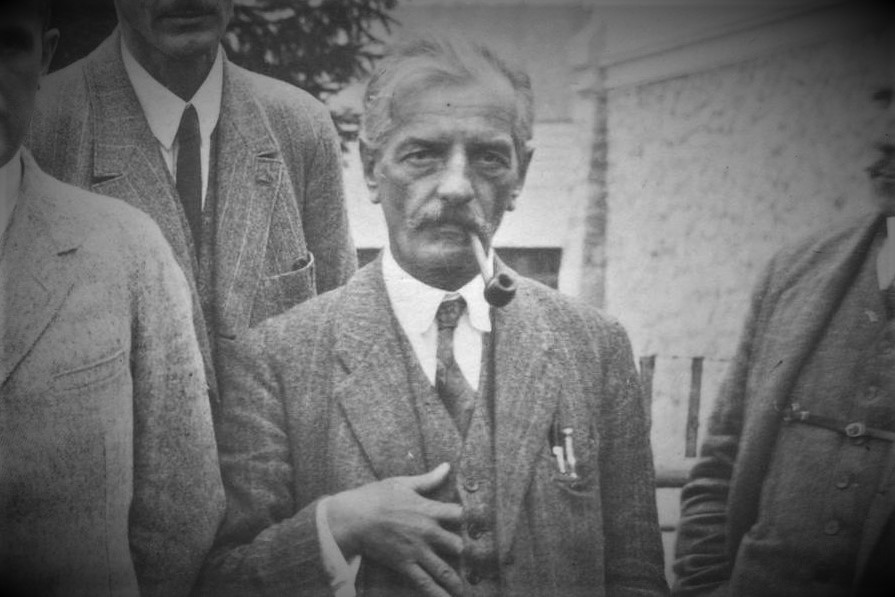 niski starszy mężczyzna, ujęcie od pasa w górę,ma fajkę w ustach dłoń trzuma na torsie