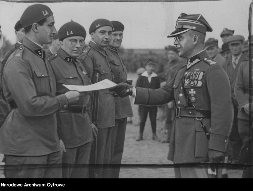 mężczyzna w mundurze z medalami wręcza niższym rangą żołnierzom w beretach dokument