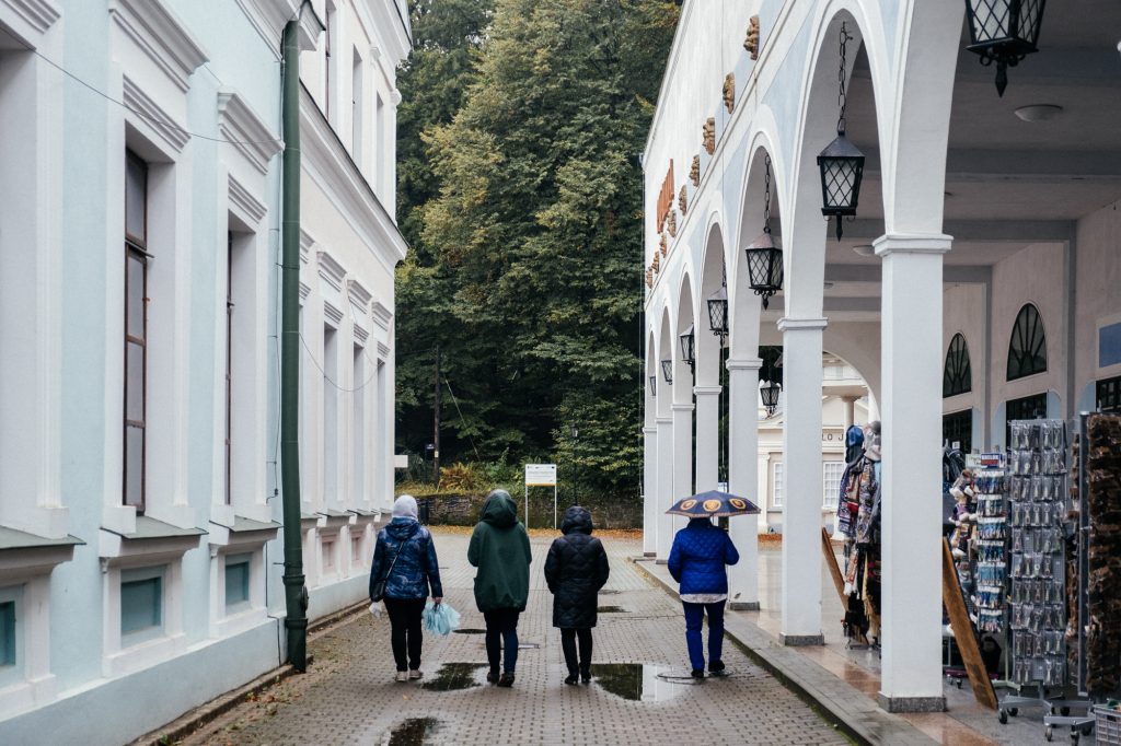 4 osoby idące przejściem pomiędzy dwoma białymi budynkami w deszczową pogodę