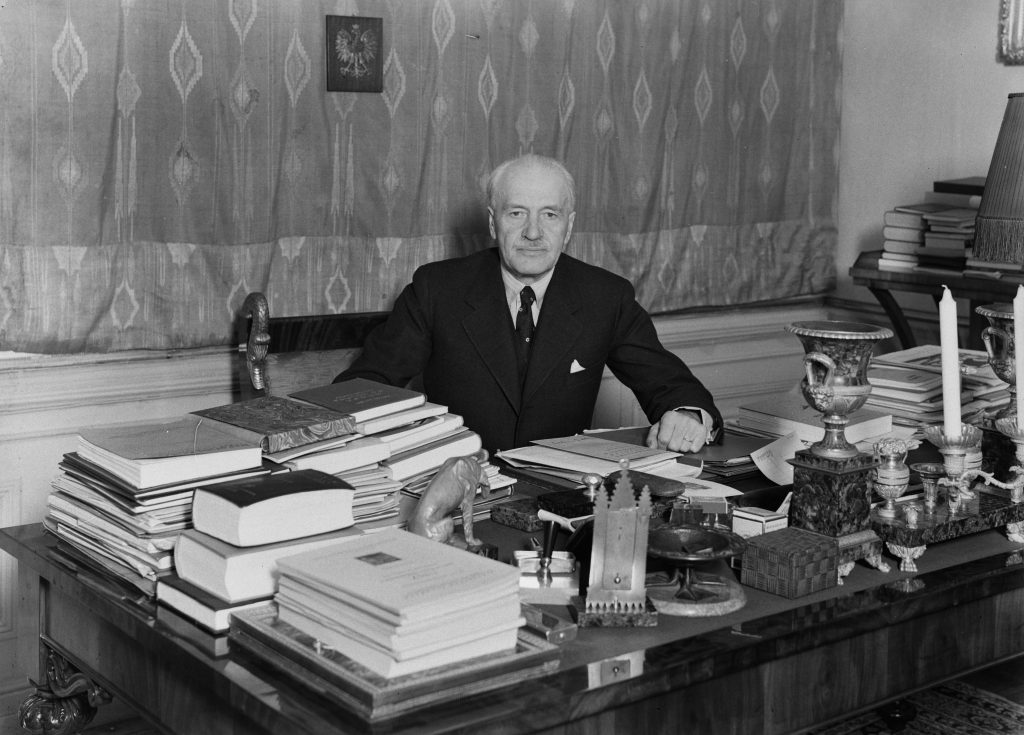 starszy mężczyzna w garniturze za biurkiem obłożonym rzeczami