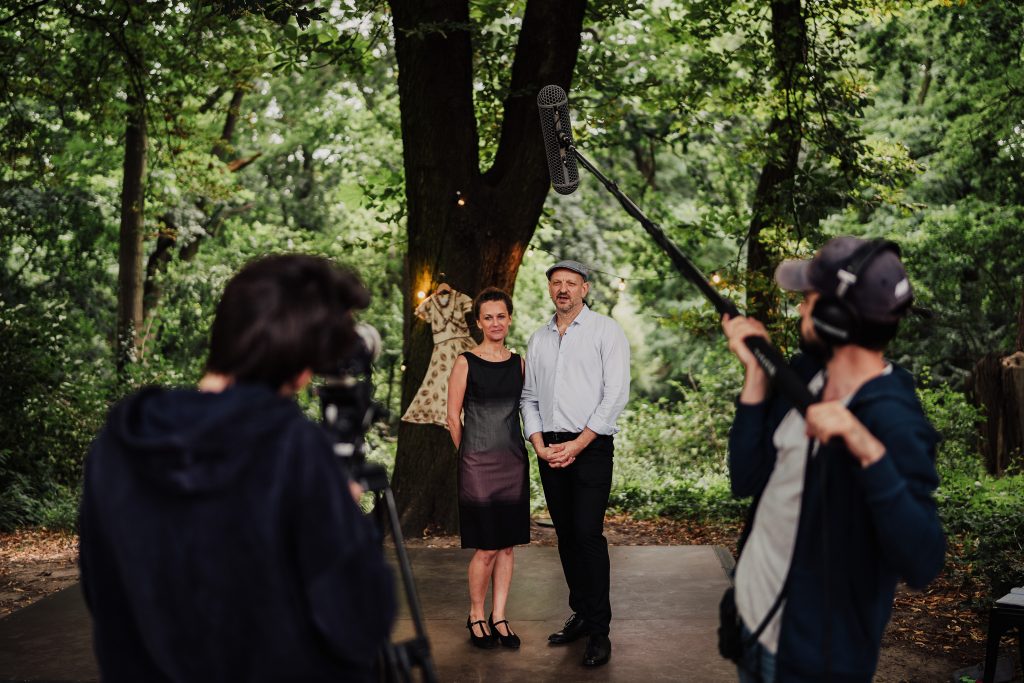 w centrum para, która prowadzi kurs, przed nimi operator kamery i operator mikrofonu realizujący nagranie, w tle drzewa i zieleń ogródka Niepodległa. Miejsce Spotkań