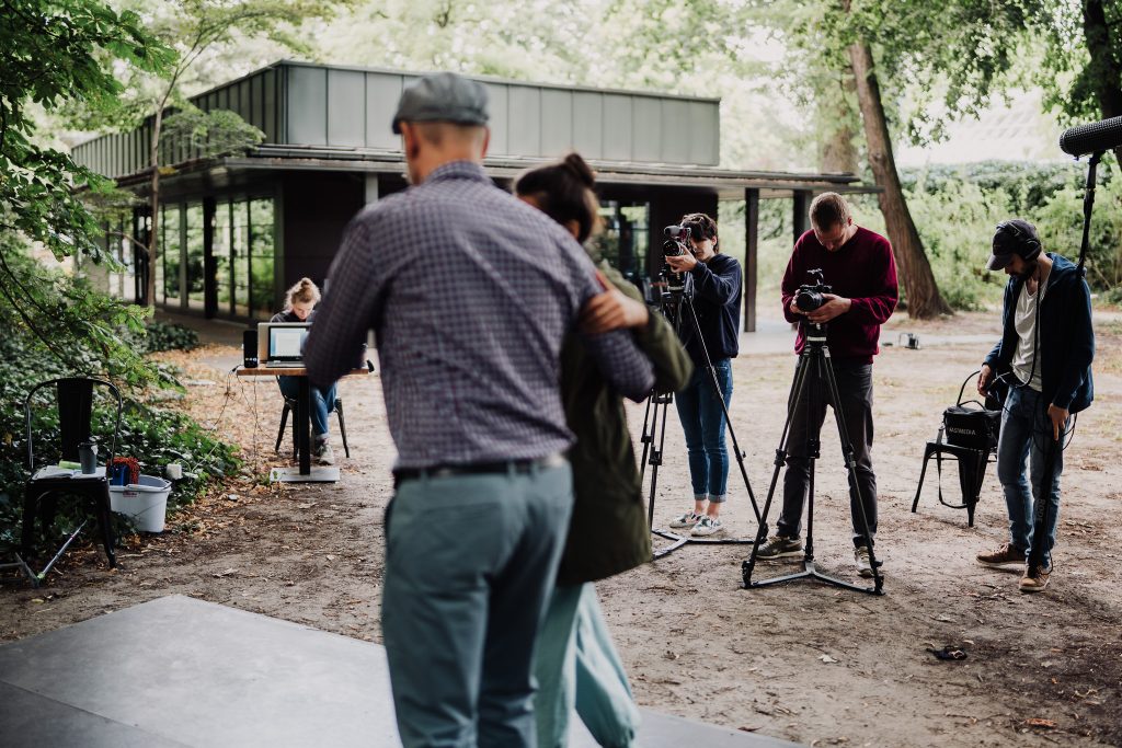 na pierwszym planie odwrócona tyłem tańcząca para, za nimi ekipa filmowo-dźwiękowa, w tle budynek niepodległa miejsce spotkań