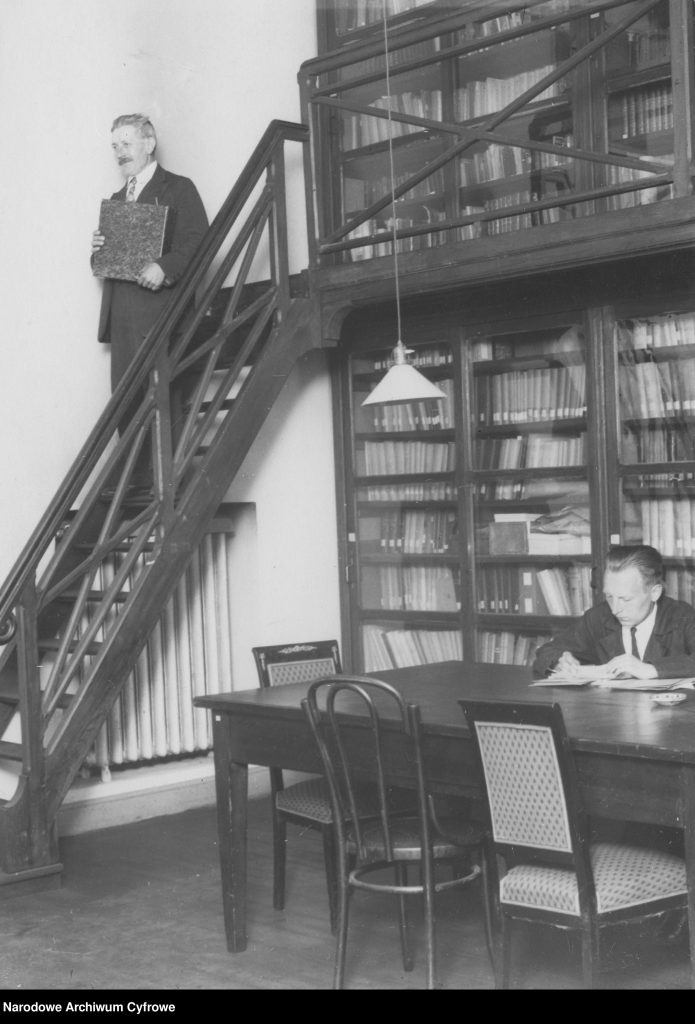 fragment pomieszczenia ze stołem, regałami i antresolą a także dwoma mężczyznami przy pracy naukowej