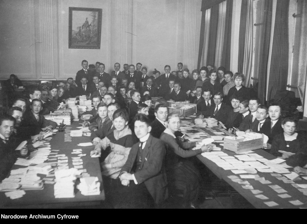 ludzie usadzeni przy długich stołach zarzuconych kartkami papieru