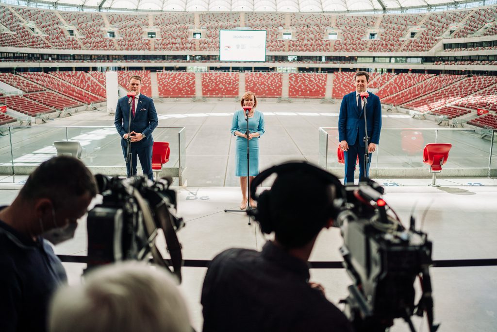 trzy elegancko ubrane osoby przed mikrofonami, w tle trybuny PGE Narodowego, na pierwszym planie kamery