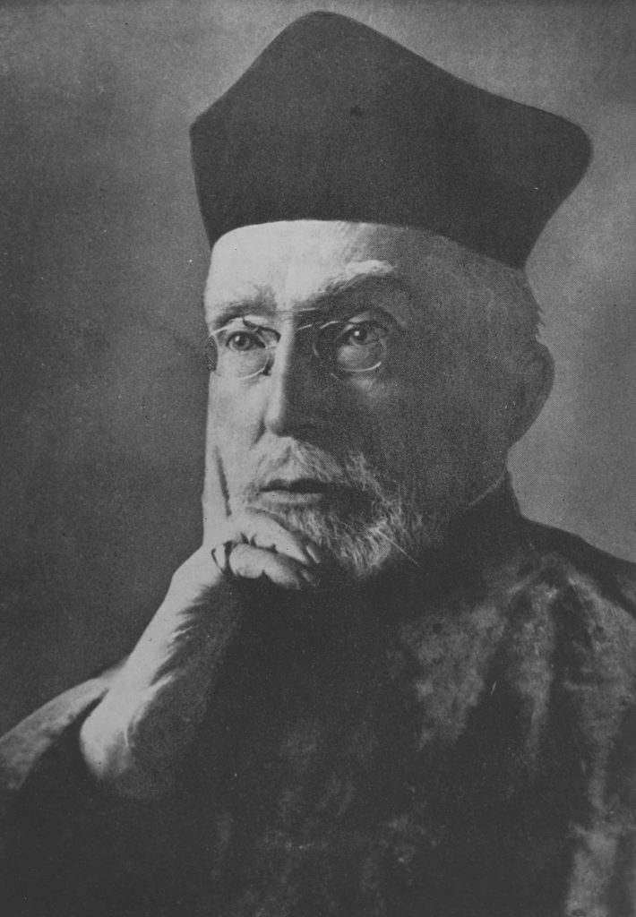 mężczyzna w todzie i czapce profesorskiej w okularach pozujący do zdjęcia w zamyślonej pozie z brodą i policzkiem opartym o dłoń