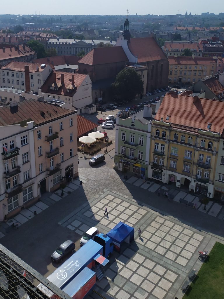 widok z góry na rozstawiony samochod stojący na rynku pomiędzy kamieniczkami, w tle kościół i zabudowa miejska Kalisza