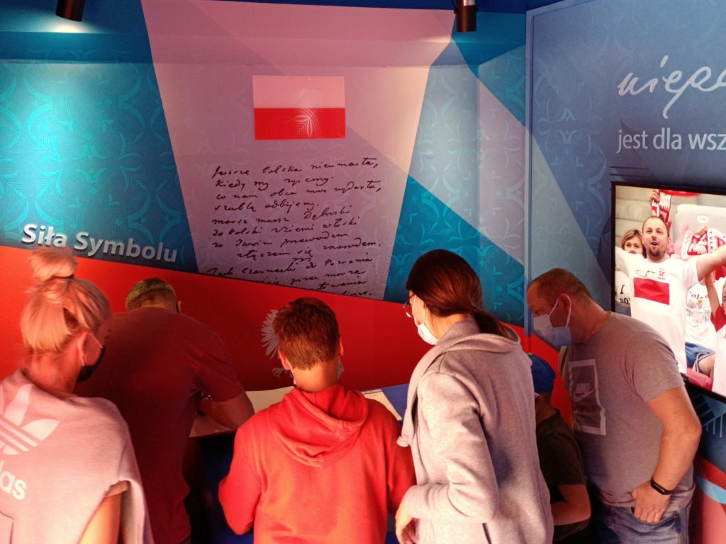 pięcioro ludzi pochyla się nad księgą w której jeden mężczyzna dokonuje wpisu, w tle grafiki wnętrza WTH