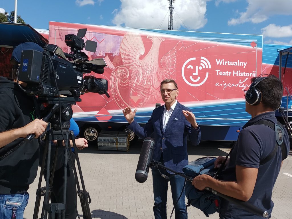 przed oklejoną ciężarówką stoi elegancki mężczyzna i opowiada do kamery i dziennikarza