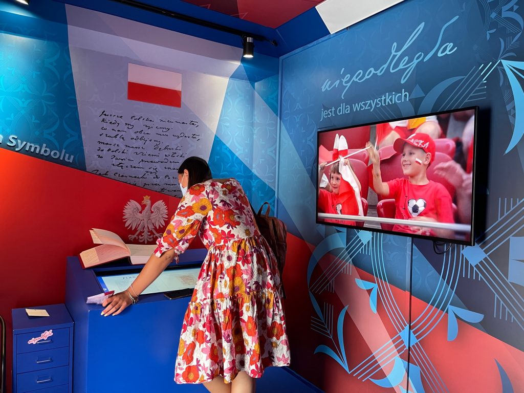 zdjęcie przedstawia kobietę w sukience oglądającą multimedialną ekspozycję w autokarze zaadaptowanym na inteaktywną przestrzeń wystawienniczą