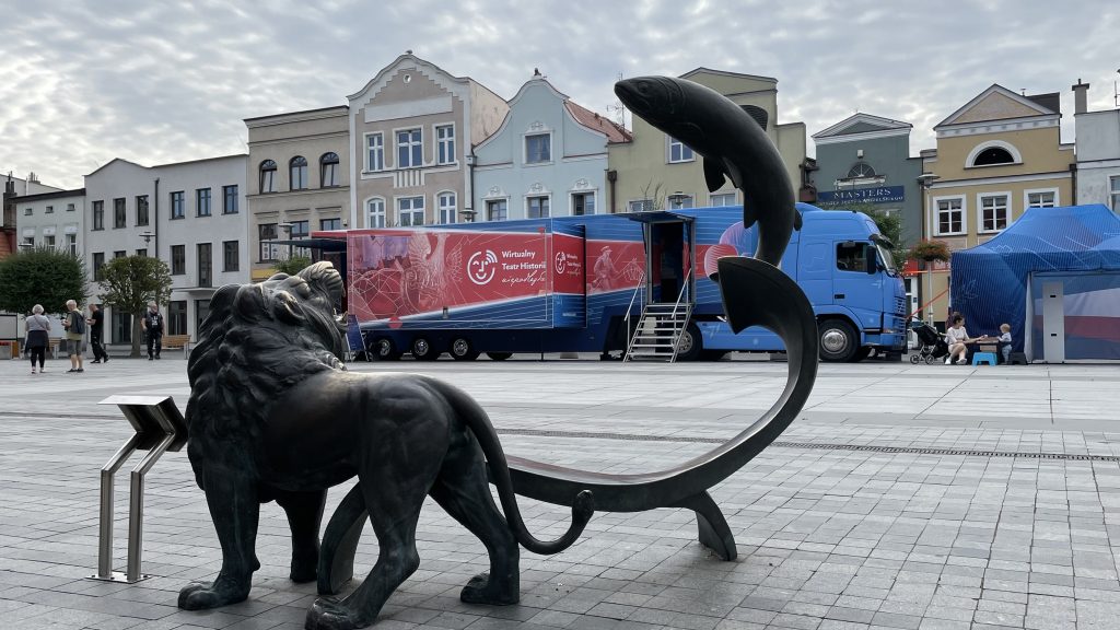 rozstawiony bus i namiot z fotobudką na rynku, na pierwszym planie pucka rzeźba 