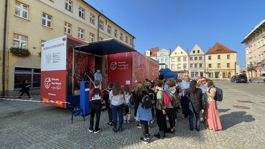 autobus z wystawą stojący na tle kamieniczek, przed wejściem tłum dzieciaków