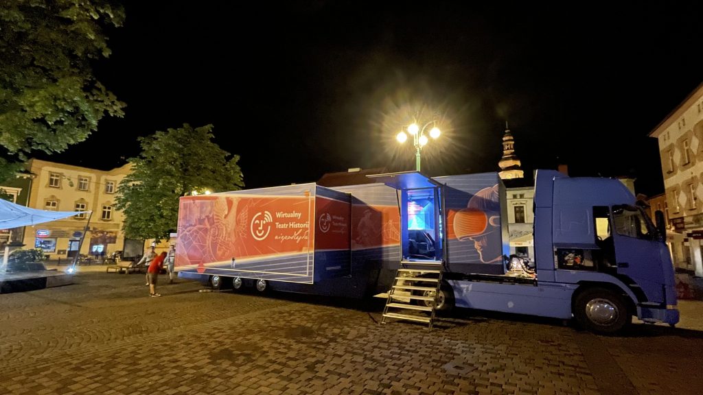 rozłożony bus na miejskim rynku nocą, w tle zabytkowa wieża