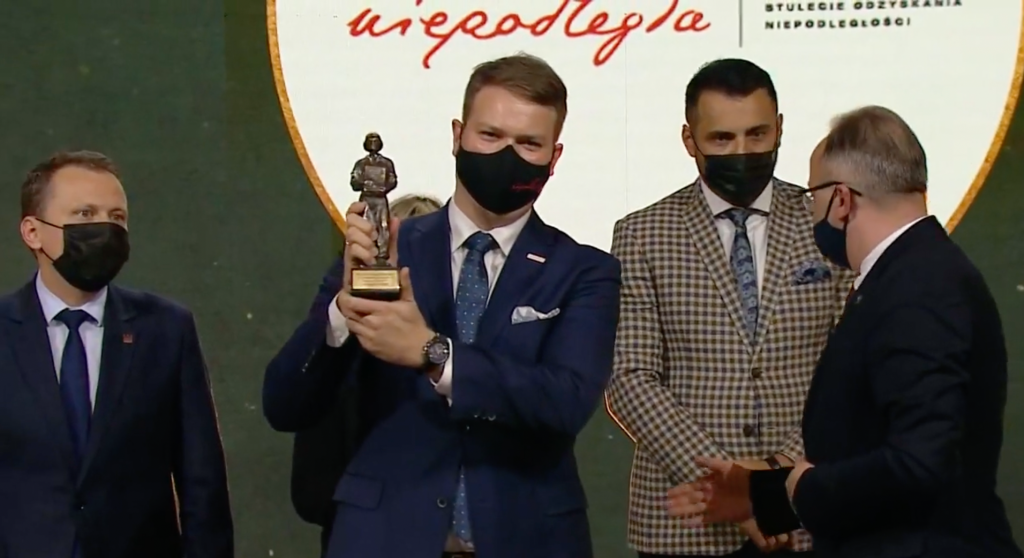 mężczyzna z uśmiechem schowanym za maseczką prezentuje nagrodę, w tle trzej inni mężczyźni