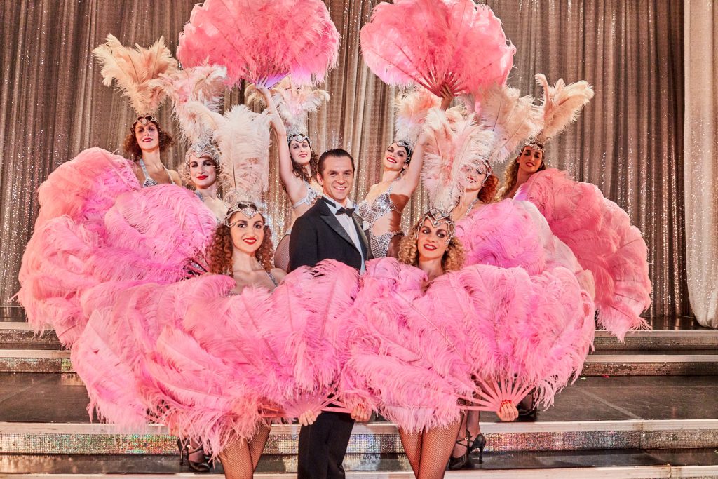 aktor we fraku otoczony przez tancerki z wielkimi pierzastymi wahlarzami