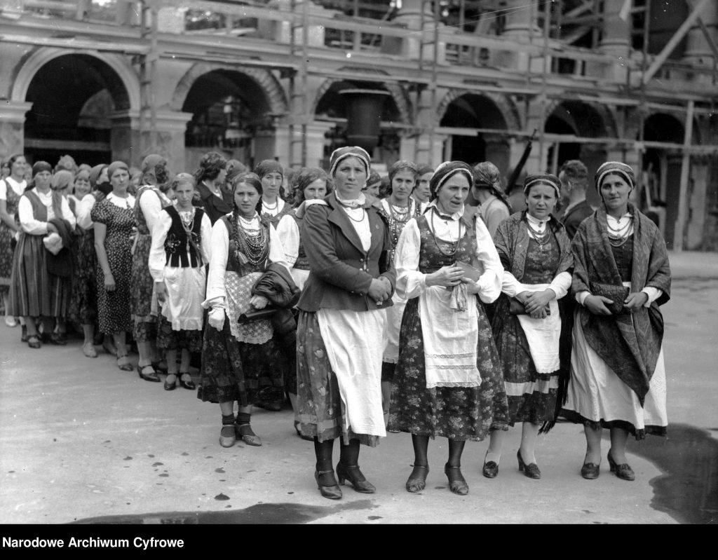 kobiety w strojach ludowych maszerują w kolumnie, w tle elementy architektury