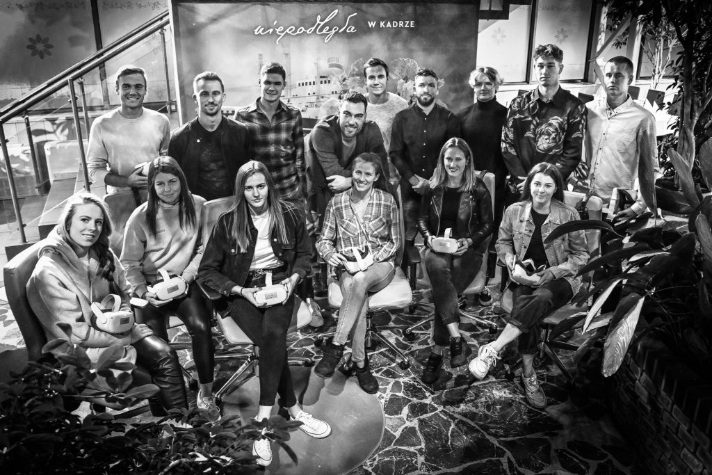 grupa ludzi pozuje na tle ścianki z logo niepodległa, osoby w pierwszym rzędzie trzymają w rękach okulusy