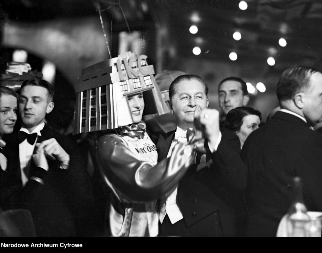 parkiet taneczny, mężczyzna w smokingu prowadzi w tańcu kobietę w trapezowym futurystycznym nakryciem głowy z napisem IKSC