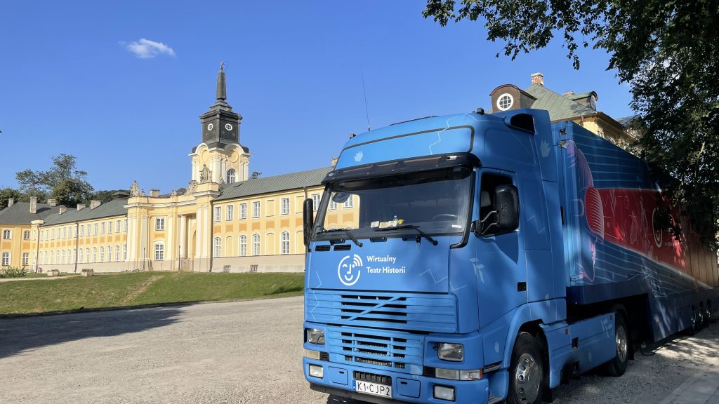 samochód Wirtualnego Teatru Historii na parkingu, w tle budynek pałacu z wieżyczką