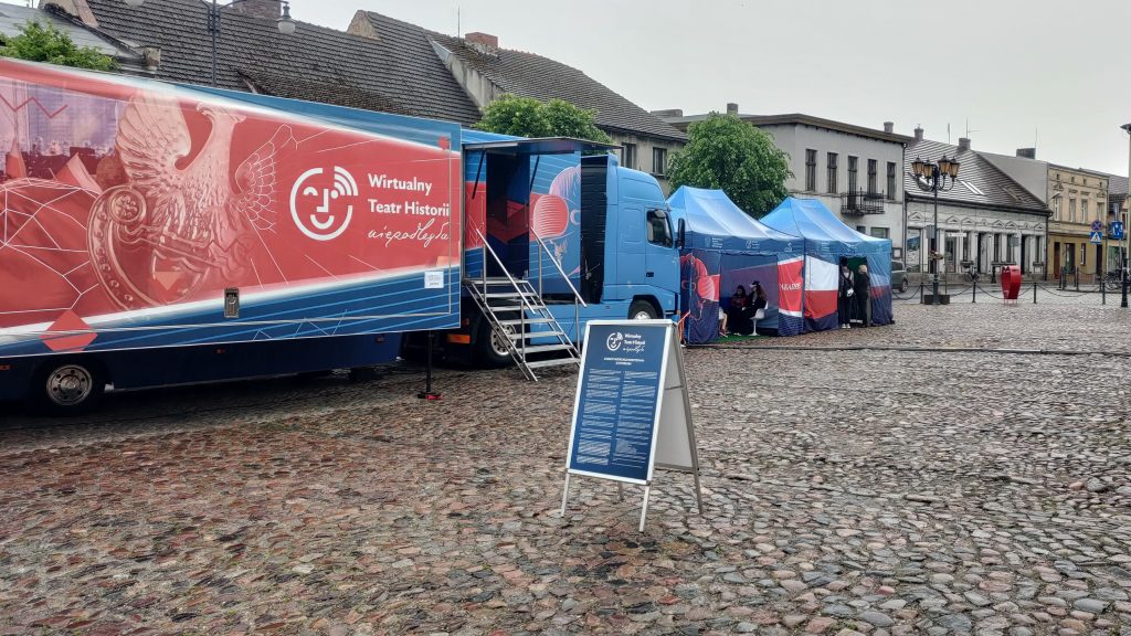 samochód Wirtualnego Teatru Historii na rynku wyłożonym kamieniami, w tle budynki pierzei