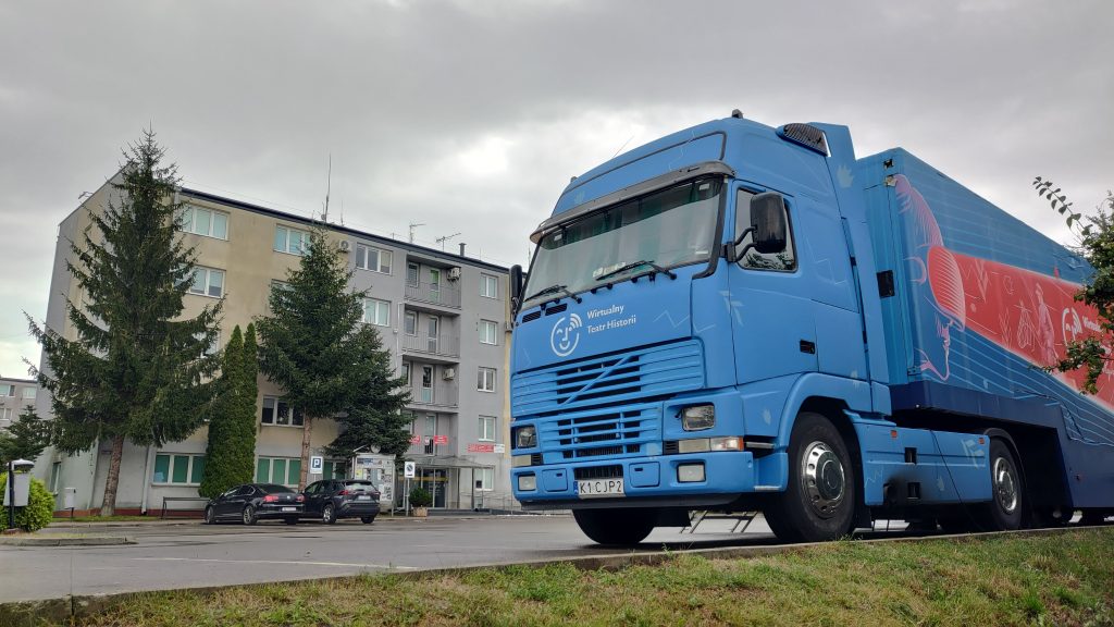 samochód Wirtualnego Teatru Historii na parkingu, w tle budynek urzędu w formie 4-piętrowego bloku