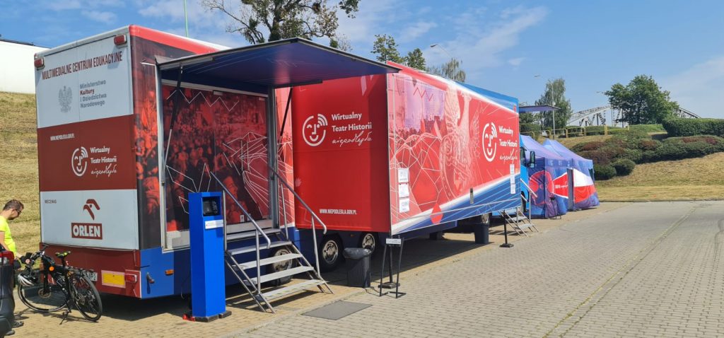 samochód Wirtualnego Teatru Historii na parkingu, w tle wał nad wisłą i za nim widoczny fragment mostu