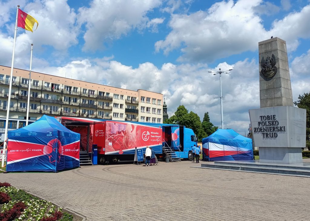 samochód Wirtualnego Teatru Historii na placu we Włocławku, za nim pierzeja z bloków, obok stoi pomnik poświęcony żołnierzom polskim