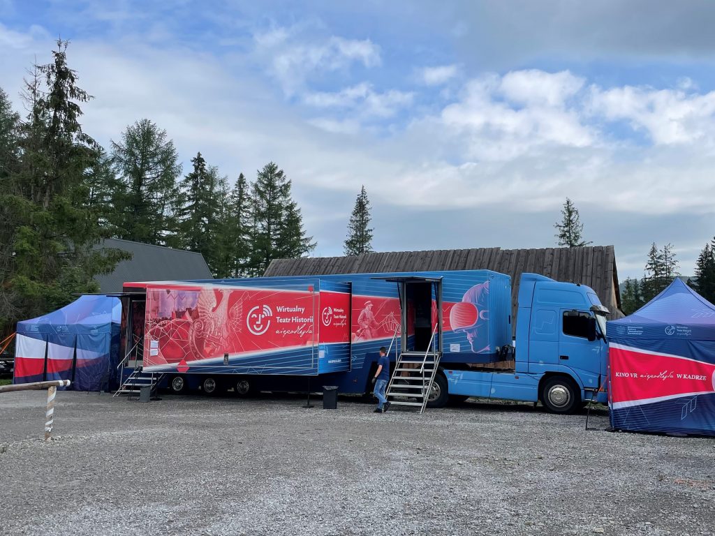 samochód Wirtualnego Teatru Historii na tle drewnianej chaty i drzew iglastych