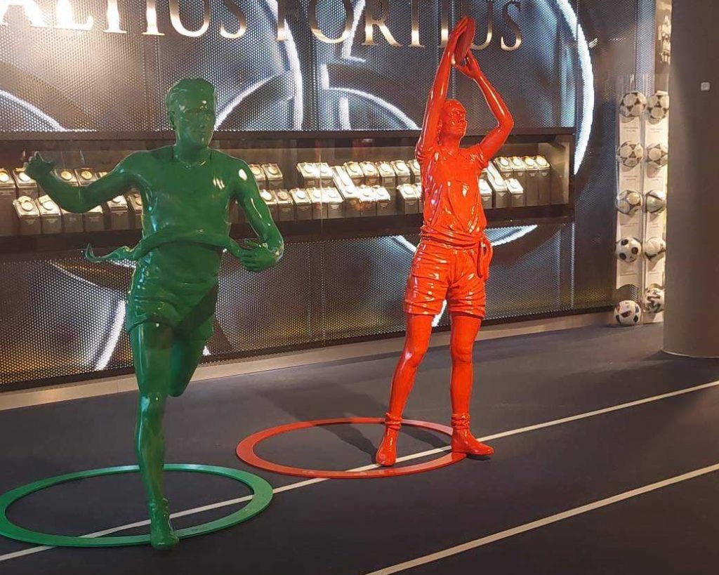 dwie rzeźby w kontrastowych kolorach ustawione na tle ekspozycji z medalami i piłkami