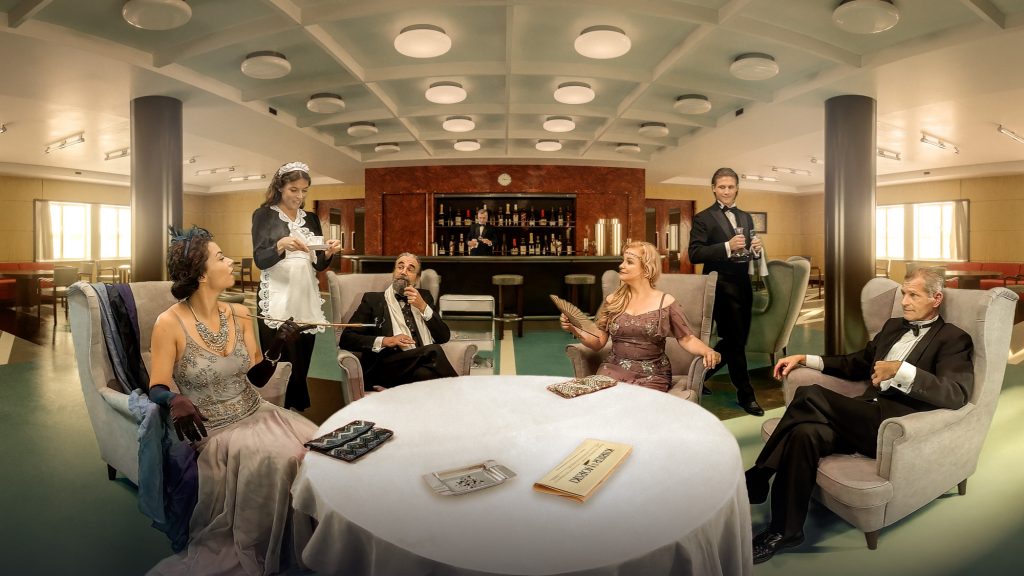 czwórka arystokratów przy stole w palarni, stoją pokojówka i młody chłopak