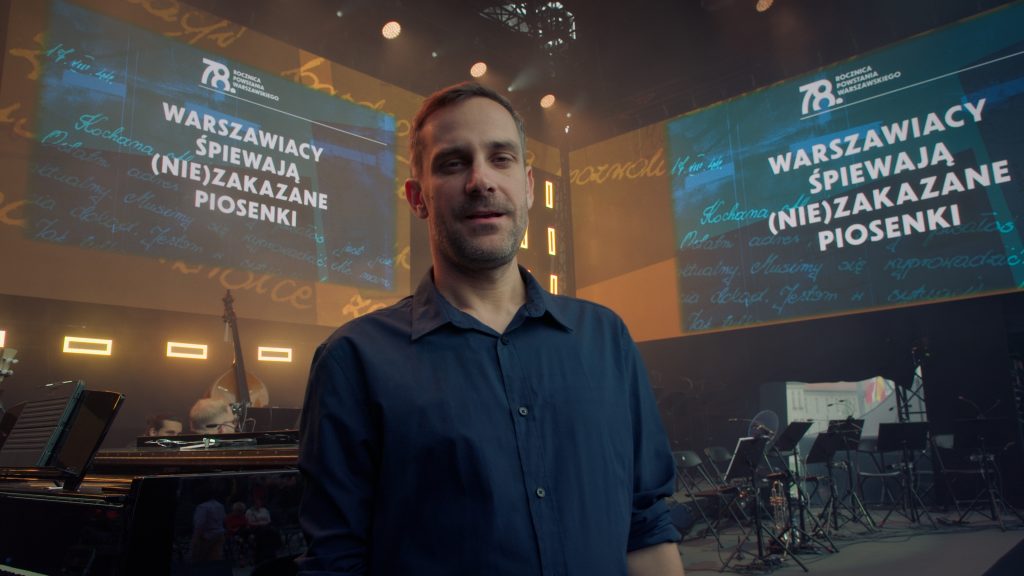 mężczyzna w koszuli, w tle ekrany z tytułem warszawiacy śpiewają niezakazane piosenki