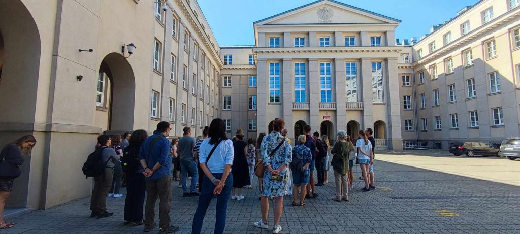 grupa ludzi stojąca na dziedzińcu, naprzeciw nich przewodnik