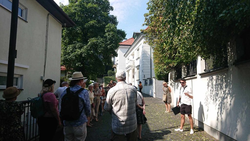 grupa ludzi stojąca w uliczce z willami, naprzeciw nich przewodnik