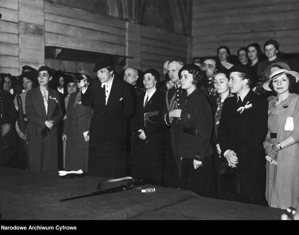 pośrodku grupy ludzi Aleksandra Piłsudska celująca z rewolweru do tarczy. przed nią stół z innym karabinkiem.