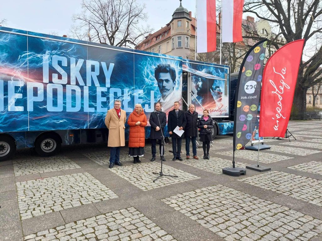 Naczepa iskry niepodległej na brukowanym placu, w tle zabudowa miejska, przed naczepą 6 oficjeli otwierających trasę