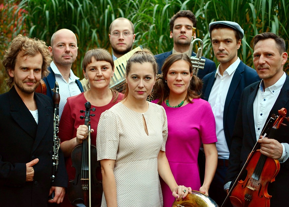 Na zdjęciu widać 9 członków zespołu Tęgie Chłopy, trzy kobiety i sześciu mężczyzn. Tło zielone.