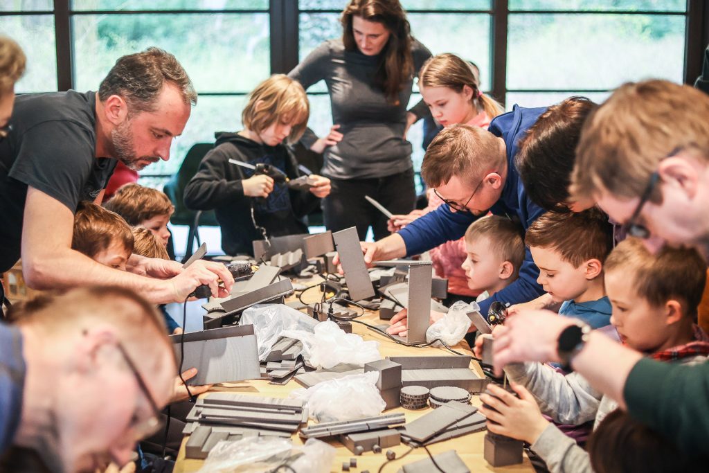 Zdjęcie przedstawia grupę dzieci i dorosłych, którzy stoją przy długim stole. Wykonują pracę plastyczną z użyciem gorącego kleju i specjalniego styropianu.